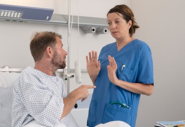 Eine Pflegerin weicht vor einem Patienten zurück, der mit dem Finger auf sie zeigt. Sie hebt abwehrend die Hände. Die Pflegerin trägt einen blauen Kittel. Der Patient ein Krankenhausnachthemd. Eine Pflegerin weicht vor einem Patienten zurück, der mit dem Finger auf sie zeigt. Sie hebt abwehrend die Hände. Die Pflegerin trägt einen blauen Kittel. Der Patient ein Krankenhausnachthemd.