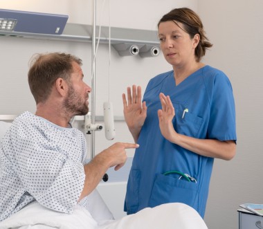 Eine Pflegerin weicht vor einem Patienten zurück, der mit dem Finger auf sie zeigt. Sie hebt abwehrend die Hände. Die Pflegerin trägt einen blauen Kittel. Der Patient ein Krankenhausnachthemd. Eine Pflegerin weicht vor einem Patienten zurück, der mit dem Finger auf sie zeigt. Sie hebt abwehrend die Hände. Die Pflegerin trägt einen blauen Kittel. Der Patient ein Krankenhausnachthemd.