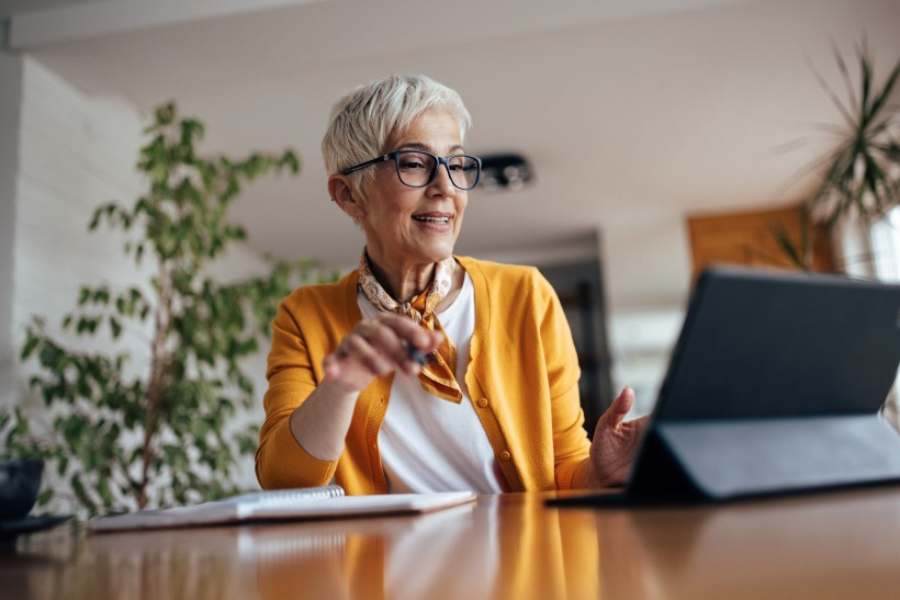 Ältere Frau mit kurzem grauen Haar, Brille und gelber Strickjacke sitzt an einem Tisch und schaut lächelnd auf ein Tablet. In der Hand hält sie einen Stift, daneben liegt ein Notizbuch. Die Umgebung wirkt wohnlich, im Hintergrund sind Pflanzen zu sehen.